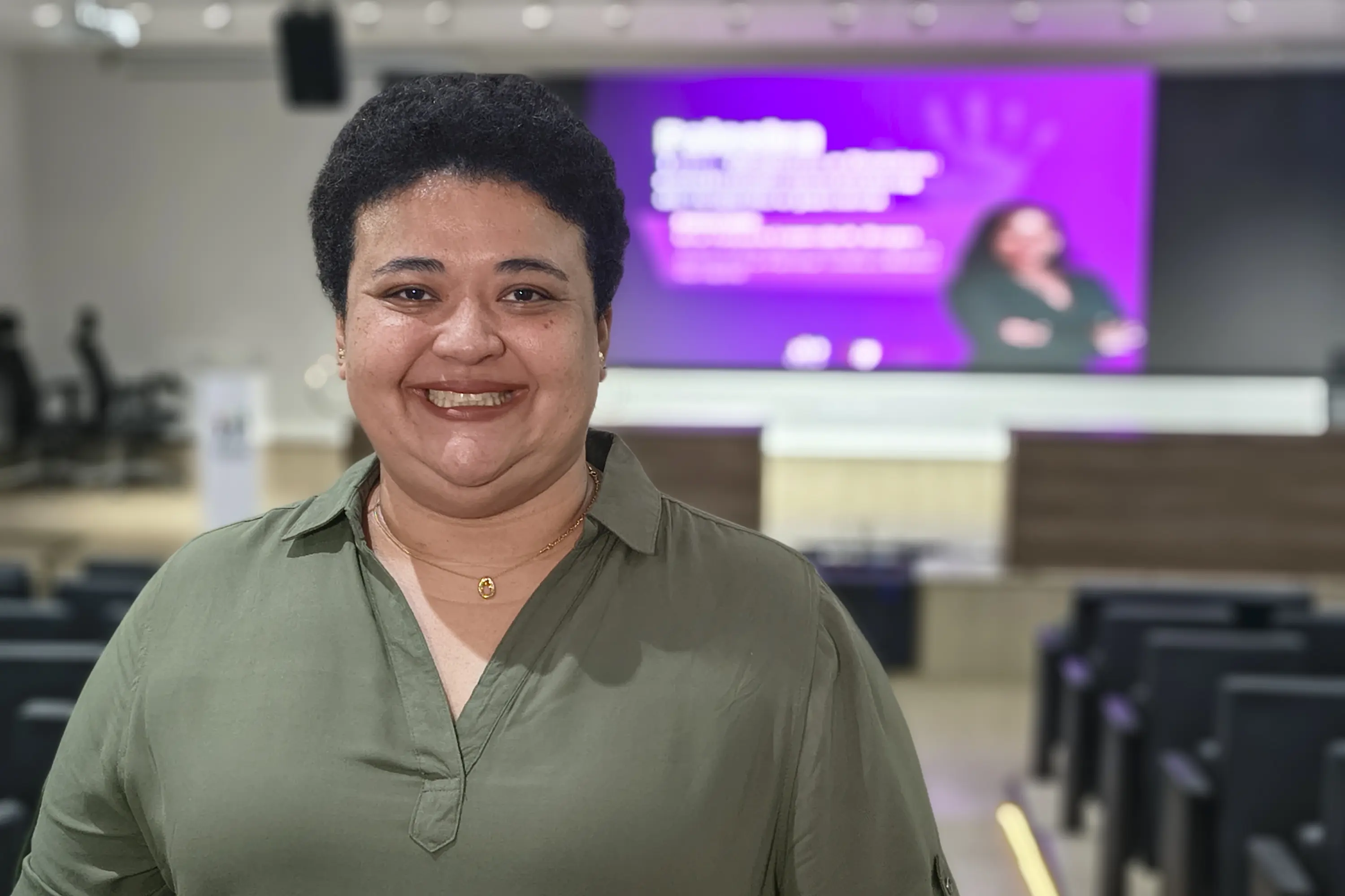 Mulher de cabelos curtos e cacheados sorri, veste blusa verde-oliva. Ao fundo, auditório com telão roxo exibindo slide e outra palestrante desfocada.