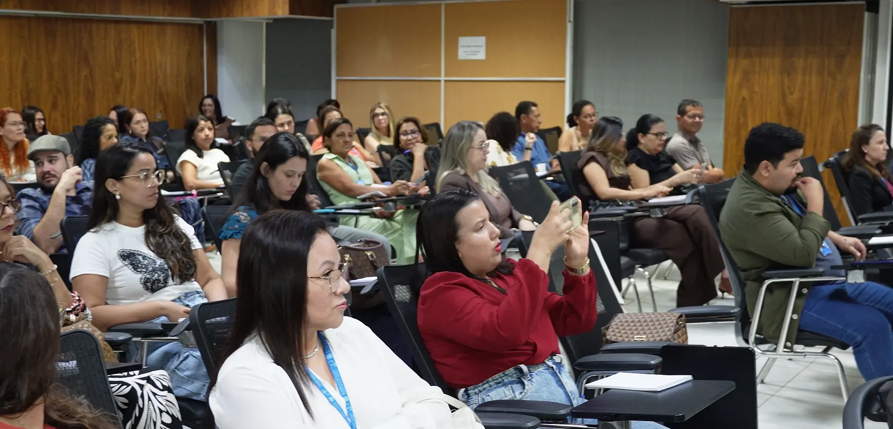Diversas pessoas sentadas em cadeiras com prancheta em um auditório. Em destaque no centro, uma mulher de blusa vermelha segura um celular horizontalmente para tirar uma foto. Outras pessoas ao redor observam atentamente.