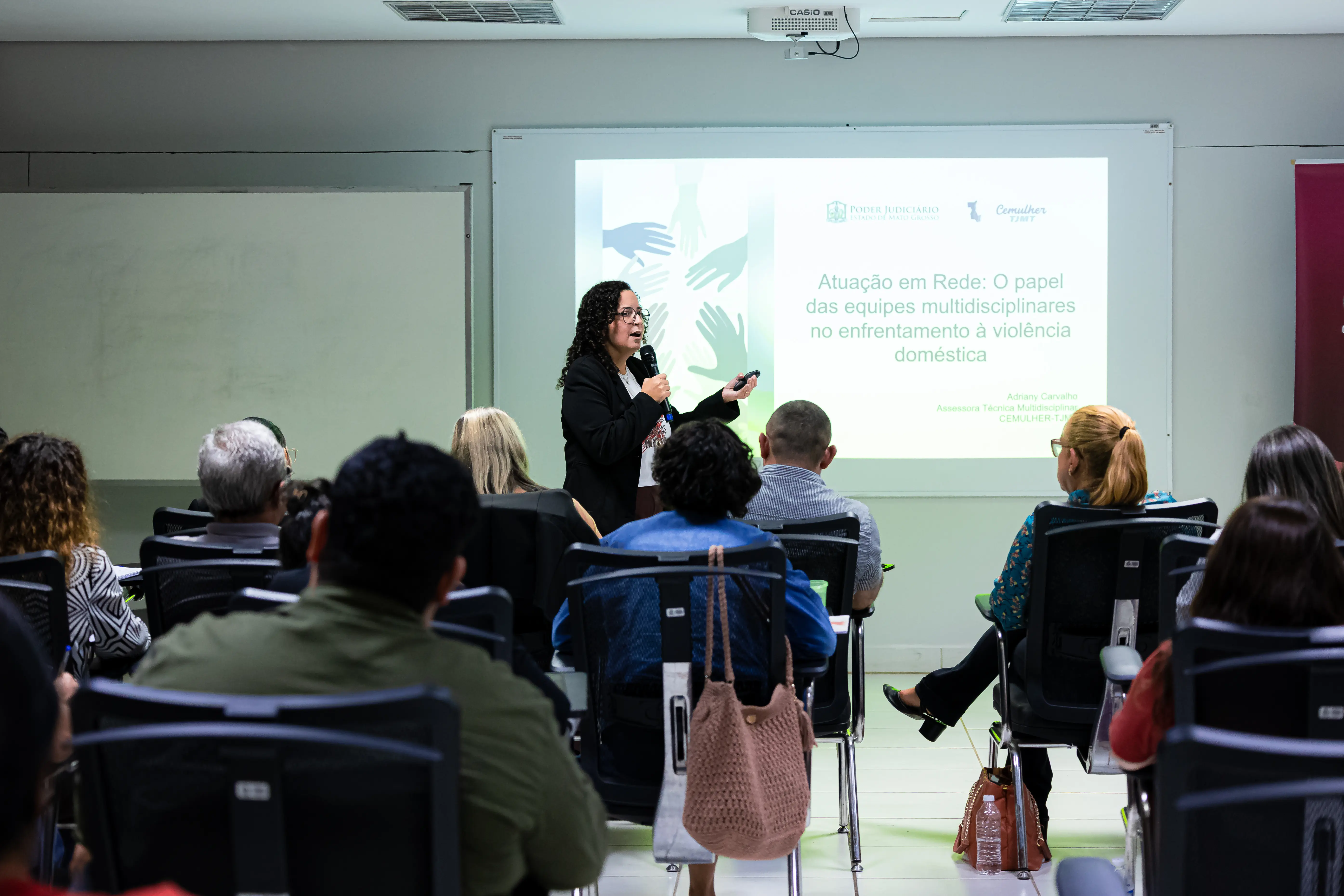 Participantes acompanham a palestra da assessora técnica multidisciplinar da Cemulher-MT, Adriany Sthefany de Carvalho, durante a capacitação das equipes multidisciplinares das Varas Especializadas de Violência Doméstica e Familiar contra a Mulher. Ao fundo, um telão exibe o tema da apresentação.