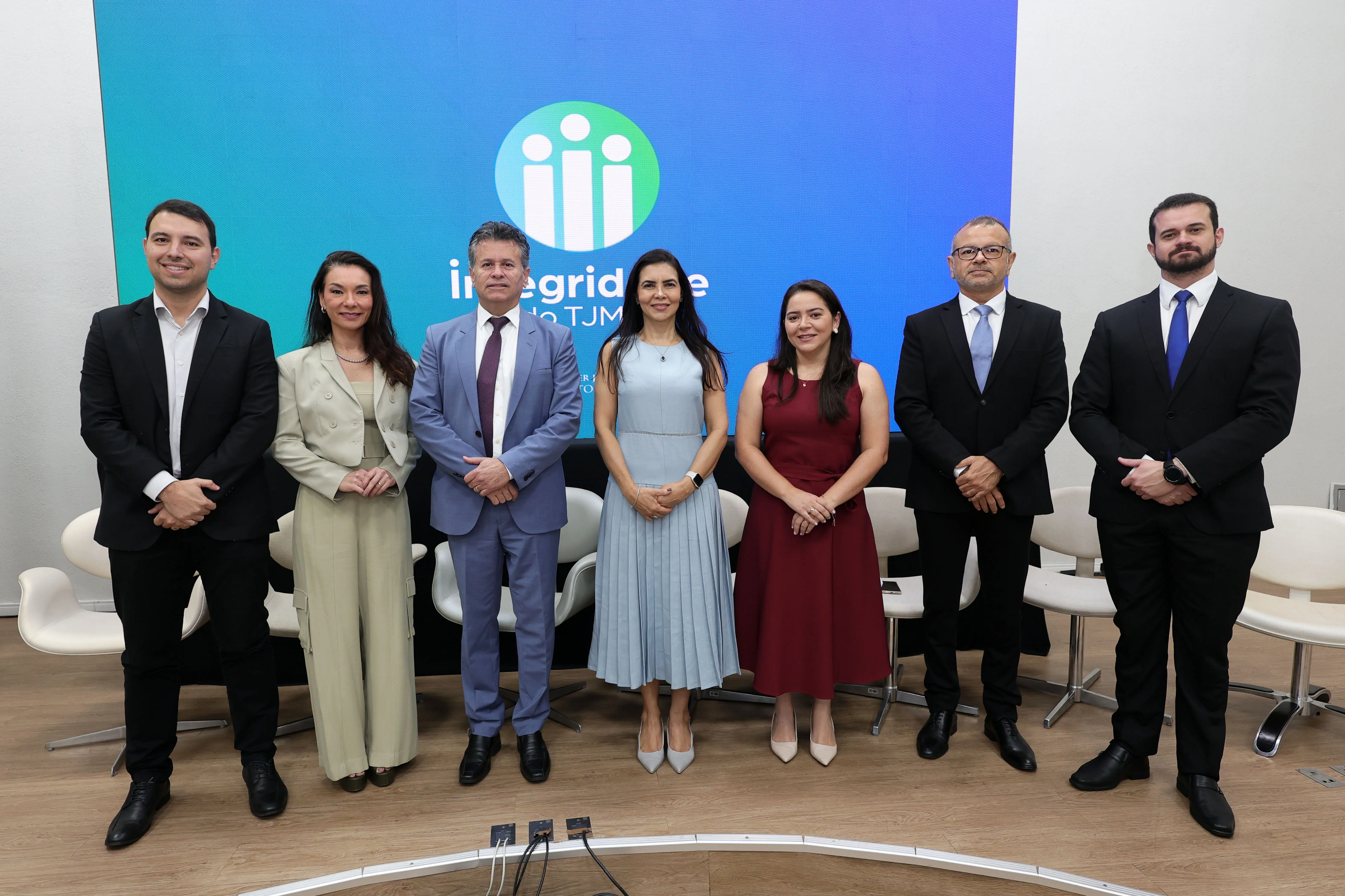 Sete pessoas em trajes formais posam alinhadas no palco de um auditório, em frente a poltronas brancas e a um telão azul com o logotipo