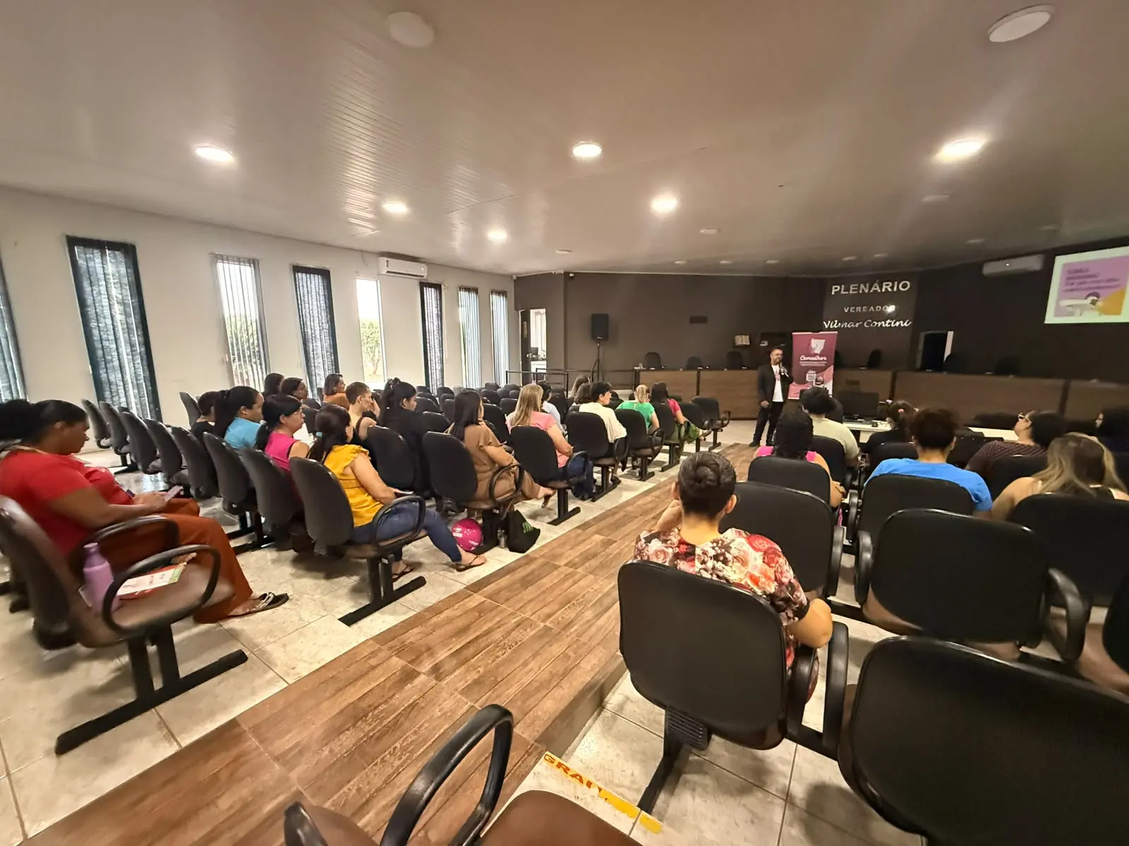 Plenário da Câmara Municipal com diversas mulheres assistindo a uma palestra. O palestrante está em pé na frente, e uma tela projeta conteúdo ao lado direito.