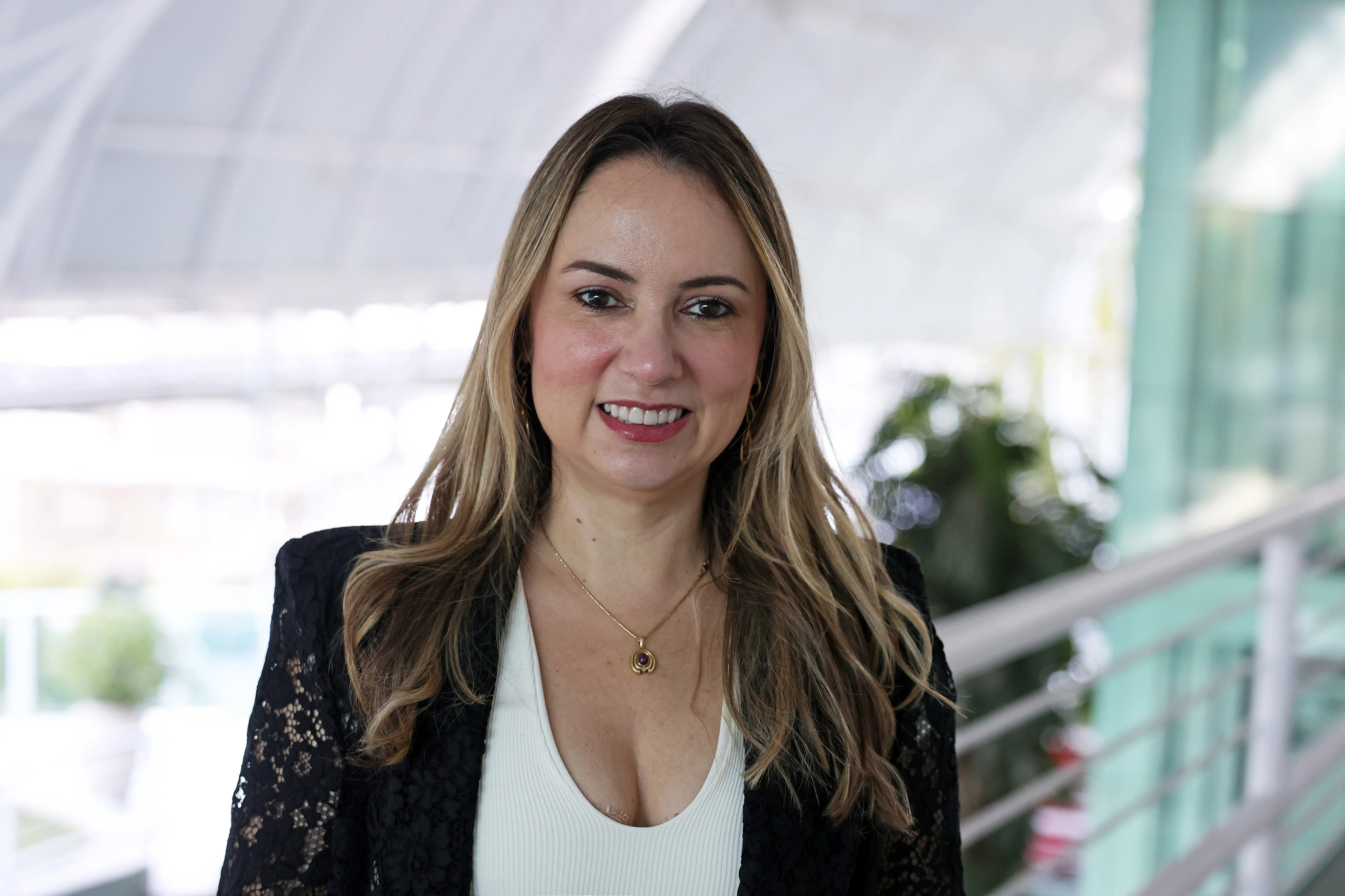 Mulher de cabelos longos e loiros, veste blazer preto rendado sobre blusa branca e colar dourado, sorri em ambiente iluminado. Ao fundo, plantas e estrutura de vidro