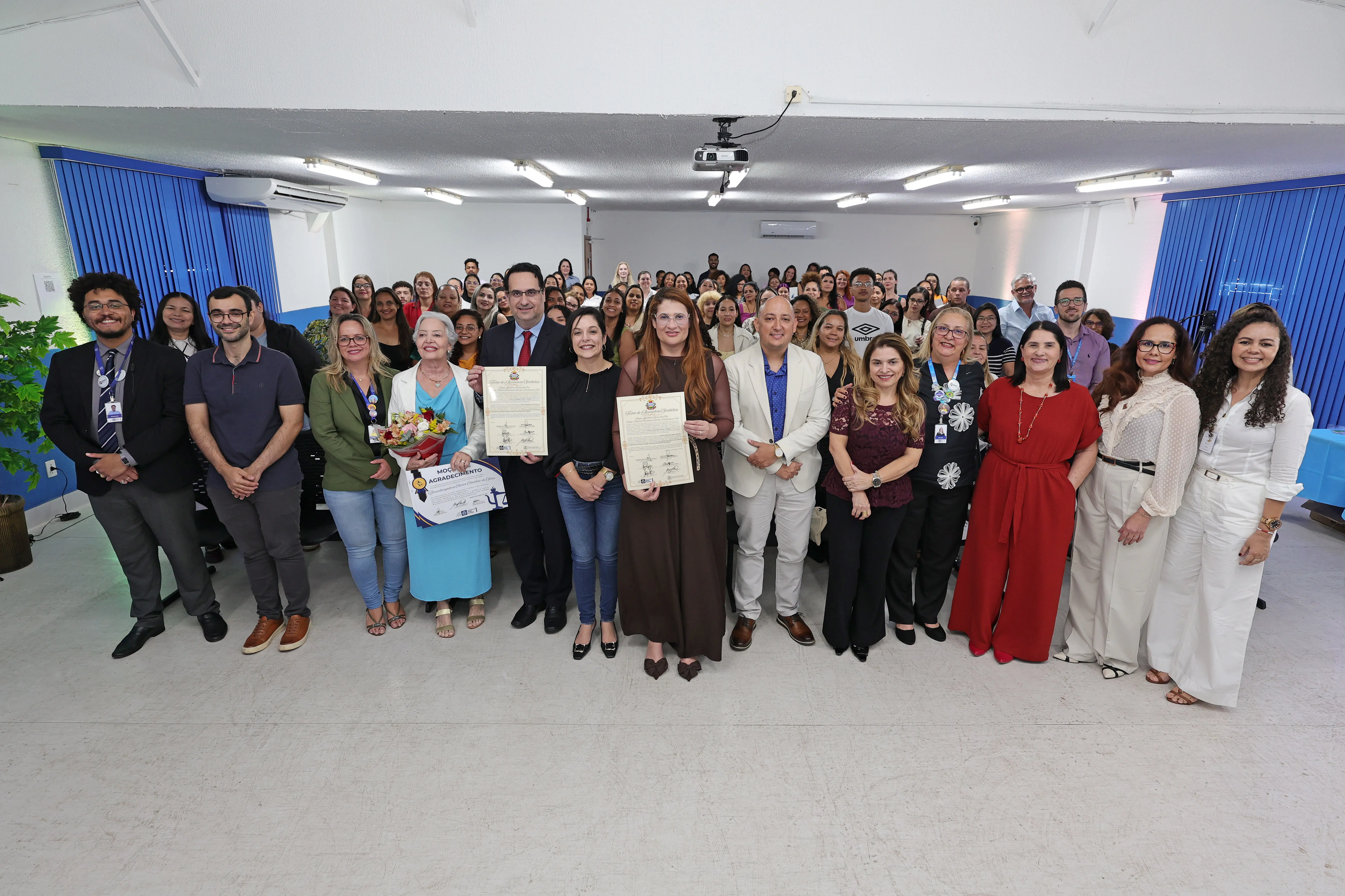 Grupo de facilitadores e autoridades posa para foto em auditório. À frente, um homem de terno escuro e uma mulher de vestido marrom exibem certificados emoldurados recebidos durante o evento.